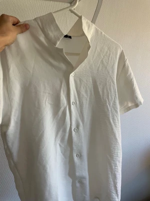 Vit kortärmad skjorta med knappar (oversize) - Stilren vit skjorta med korta ärmar och knäppning framtill. Skjortan har en klassisk krage och är tillverkad i ett lätt material som passar perfekt till varmare dagar. Enkel och clean design som funkar till många olika stilar.