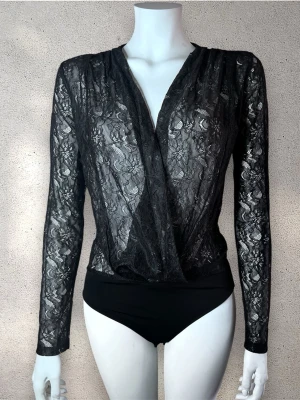 Zara spetsbody med omlott - Snygg svart body i transparent spets med omlott framtill och långa ärmar. Bodyn har ett klassiskt snitt och är tillverkad i ett spetsmaterial som ger en elegant look. Perfekt för dig som gillar trendiga och stilrena underkläder. #zara
