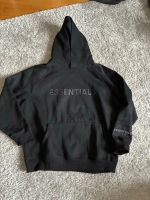 Svart Essentials hoodie med huva - Säljer en svart hoodie från Essentials med stor huva och rymlig magficka. Hoodien har tryckt logotyp på bröstet och ärmen. Materialet är mjukt och bekvämt, perfekt för en avslappnad look. Passar dig som gillar streetwear och enkel stil.