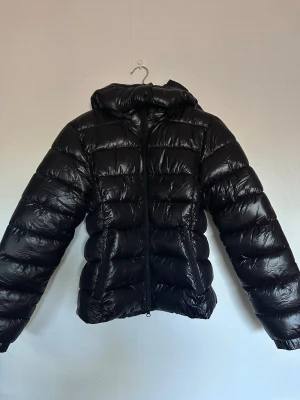 Svart pufferjacka med huva - Säljer en svart pufferjacka med glansig finish och huva. Jackan har dragkedja framtill och quiltad design med breda kanaler. Perfekt för kalla dagar och ger en cool look. Materialet är syntet och jackan är lätt vadderad.