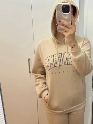 Beige hoodie med tryck 'ANGELS' - Säljer en beige hoodie med huva och dragsnören med byxor samma färg . På bröstet står det 'ANGELS CALIFORNIA' i stora bokstäver. Hoodien har en klassisk känguruficka och ribbade muddar. Perfekt för en chill och avslappnad stil.