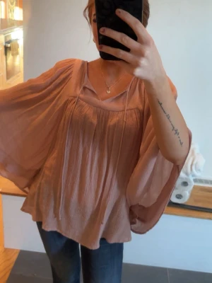 Ljusrosa flowy blus med vida ärmar - Superfin ljusrosa blus med flowy och luftig passform. Blusen har vida, långa ärmar och ett snyggt fall i tyget. Materialet är tunt och lätt, perfekt för en trendig och avslappnad look. Snygga detaljer vid halsringningen ger extra stilpoäng.