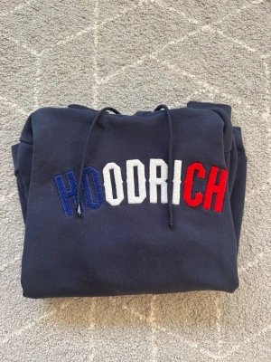 Mörkblå HOODRICH hoodie med brodyr - Snygg mörkblå hoodie från HOODRICH med broderad logga i blått, vitt och rött på bröstet. Klassisk känguruficka framtill och justerbar huva med snören. Perfekt för en avslappnad streetwear-look.