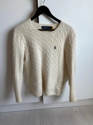 Vit kabelstickad tröja Polo Ralph Lauren  - Vit kabelstickad tröja från Polo Ralph Lauren med rund hals och broderad logotyp på bröstet. Tröjan är gjord i mjukt material och har ribbade muddar vid ärmslut och nederkant. Perfekt för dig som gillar klassisk och stilren design. Passar en S skulle jag säga. 