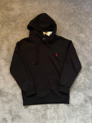 Svart hoodie från Polo Ralph Lauren - Svart hoodie med huva och dragsnören från Polo Ralph Lauren. Klassisk design med röd broderad logotyp på bröstet och en stor magficka. Mjukt material som är perfekt för en avslappnad look.