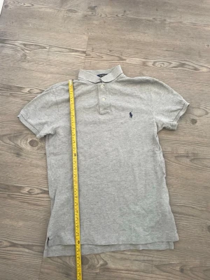 Ralph Lauren pike - Klassisk grå pikétröja från Ralph Lauren med korta ärmar och krage. Tröjan har två knappar vid halsen och den ikoniska lilla broderade loggan på bröstet. 