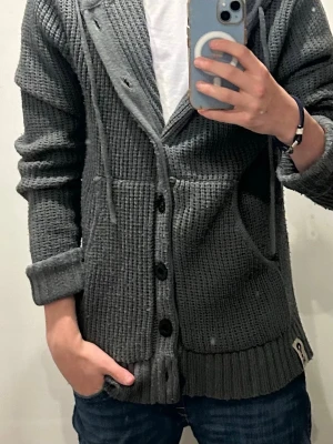 Grå stickad kofta med huva - Säljer en grå stickad kofta med huva och stora knappar framtill. Koftan har en grovstickad struktur, två fickor och ribbade muddar. Perfekt för lager-på-lager och chill stil. Passar dig som gillar avslappnat och mysigt.