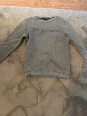 Ljusgrå sweatshirt från Sail Racing - Ljusgrå sweatshirt med rund hals och tryckt Sail Racing-logga på bröstet. Tröjan har långa ärmar och ribbade muddar vid hals, ärmslut och nederkant. Perfekt för en avslappnad och sportig stil.