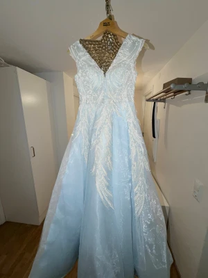 Ljusblå långklänning med pärlor - Magisk ljusblå långklänning med v-ringning och vackra vita broderier samt pärldetaljer över hela klänningen. Klänningen har ärmlös design och ett elegant, svepande fall som ger en sagolik känsla. Perfekt för bal eller speciella tillfällen.