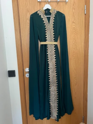 Mörkgrön långklänning med gulddekor - Elegant mörkgrön långklänning  med långa ärmar och detaljerad gulddekor längs hela framsidan och kragen. Klänningen har ett matchande guldfärgat bälte i midjan och v-ringad hals. Perfekt för speciella tillfällen.