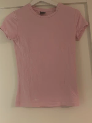 Ljusrosa t-shirt från Gina Tricot - En enkel och stilren ljusrosa t-shirt från Gina Tricot. Modellen är klassisk med rund halsringning och korta ärmar. Tillverkad i mjuk bomull som känns skön mot huden. Perfekt basplagg att matcha med allt. Är aldrig använd