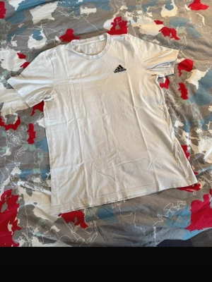 Vit Adidas t-shirt med logga - Klassisk vit t-shirt från Adidas med svart logga på bröstet. T-shirten har rund halsringning och korta ärmar. Tillverkad i mjuk bomull för en skön känsla och enkel stil.
