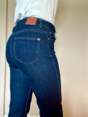 Mörkblå jeans med hög midja från Zara. Lite utsvängda  - Snygga mörkblå jeans med hög midja och klassiska bakfickor. Jeansen har kontrastsömmar och en läderpatch i midjan. Perfekta för dig som gillar en tight passform och stilren look.