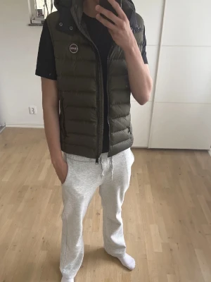 Olivgrön dunväst från Colmar - Snygg olivgrön dunväst från Colmar med svart huva och dragkedja framtill. Västen har två fickor med dragkedja och en rund logotyp på bröstet. Perfekt för lager-på-lager och streetstyle.