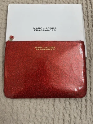 Röd glittrig necessär eller handväska från Marc Jacobs - Snygg röd glittrig clutch från Marc Jacobs Fragrances med guldtext på framsidan. Väskan har en enkel rektangulär form och stängs med dragkedja. Perfekt för att bära småsaker och ge din outfit extra bling.
