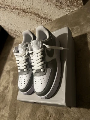 Nike Air Force 1 Low vit/grå - Nike Air Force 1 Low sneakers i vitt och grått läder. Klassisk siluett med perforerad tåbox, vita skosnören och den ikoniska Swoosh-loggan på sidan. Tjock vit sula och bekväm vadderad krage. Perfekta för en clean och stilren look.