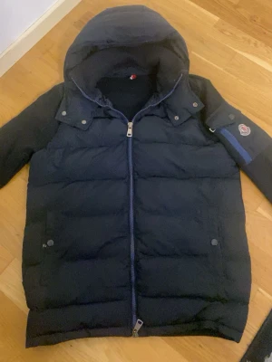 Navy blue Moncler cardigan - Snygg navy blue cardigan från Moncler med huva och dragkedja framtill. Jackan har två framfickor med knappar och en ärmficka med blå dragkedja och Moncler-logga. Tillverkad i vadderat material som håller dig varm under kalla dagar.