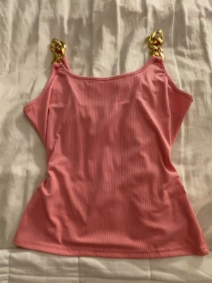 Rosa ribbad linnetopp med guldkedjor - Trendig rosa ribbad linnetopp med breda guldfärgade kedjor som axelband. Toppen är ärmlös och har en croppad passform som ger en cool och modern look. Perfekt för dig som vill sticka ut lite extra.