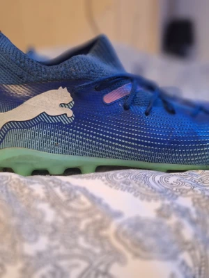 Blåa Puma fotbollsskor - Säljer ett par blåa fotbollsskor från Puma med vit logga på sidan och ljusgrön sula. Skorna har en strömlinjeformad design och är tillverkade i syntetmaterial med snörning och låg profil. Perfekta för dig som vill ha stil och komfort på planen.