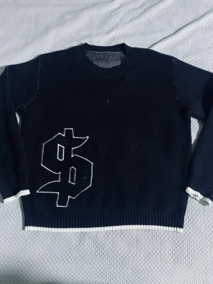 Svart stickad tröja med dollartecken - Cool svart stickad tröja med ett stort vitt dollartecken broderat på framsidan. Tröjan har rund hals, långa ärmar och vita detaljer vid ärmslut och nederkant. Perfekt för dig som gillar streetwear och vill sticka ut.