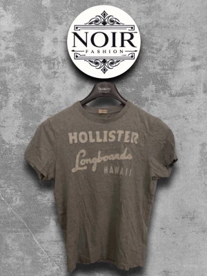 Grå Hollister t-shirt med tryck - Säljer en grå t-shirt från Hollister med texten 'Hollister Longboards Hawaii' tryckt på bröstet. Klassisk rund hals och korta ärmar. Perfekt för en avslappnad och cool stil.