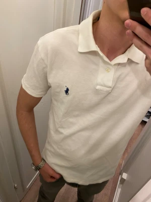Vit Piké från Ralph Lauren (Slim Fit) - Klassisk vit pikétröja från Polo Ralph Lauren med blå logga broderad på bröstet. Tröjan har korta ärmar, krage och knappar vid halsen. Tillverkad i mjuk bomull för en bekväm och stilren look. Storlek M men passar mer som S. Snabb Leverans!