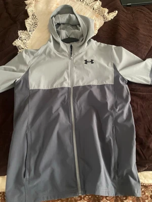 Grå regnjacka / Wind Breaker Under Armour - Snygg hoodie från Under Armour i två nyanser av grått. Dragkedja framtill, huva och två sidofickor. Tillverkad i ett mjukt och funktionellt material som passar perfekt till träning eller chill. Under Armour-logga på bröstet för en sportig vibe.