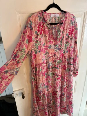 Blommig långklänning från Lil - Långklänning från by Lil i ett luftigt bomullstyg med färgglatt blommigt mönster i rosa, grönt och blått. Klänningen har långa ärmar, v-ringning med knyt och volangdetaljer vid halsen. Perfekt för sommarens/vårens alla tillfällen. Passar en stor S/M/en liten L.