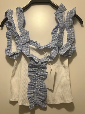 Vit ribbad topp med blå rutiga volanger - Supersöt vit ribbad linnetopp från Zara med blå och vita rutiga volangdetaljer längs axelbanden och framtill. Toppen är ärmlös och har en croppad passform som ger en trendig look. Perfekt för dig som vill sticka ut med en unik och lekfull topp.