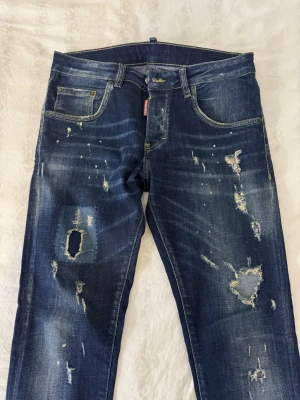 DSQUARED2 Jeans - Säljer ett par snygga Dsquared2 jeans i mörkblå tvätt med fabriks-slitningar och färgstänk. Klassisk femficksmodell med knappgylf och ikonisk röd Dsquared2-tag. 