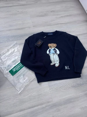 Mörkblå stickad tröja Polo Bear - XS - Mörkblå stickad tröja från Polo Ralph Lauren med den ikoniska Polo Bear-björnen broderad på framsidan. Tröjan har rund hals, långa ärmar och RL-broderi vid nederkanten. Perfekt för dig som gillar klassisk och lekfull stil. Pris kan alltid diskuteras 