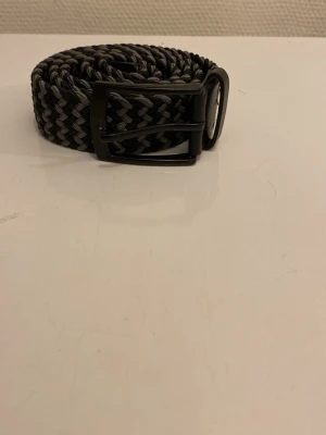 Svart och grått flätat bälte - Snyggt flätat bälte i svart och grått med metallspänne. Bältet har ett zigzag-mönster och är gjort i ett elastiskt material som gör det bekvämt att bära. Perfekt för att ge din outfit en cool touch.