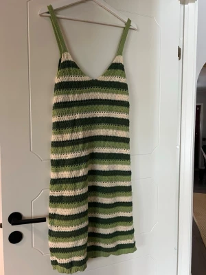 Randig stickad sommarklänning - Säljer en stickad klänning med gröna och vita ränder. Klänningen har smala axelband, v-ringad hals och ärmlös design. Perfekt för varma dagar och har en avslappnad passform. Materialet är mjukt och luftigt, vilket gör den skön att bära under sommaren.