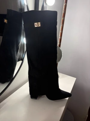 Svarta knähöga boots med gulddetalj - Säljer ett par svarta knähöga stövlar med spetsig tå och chunky klack. Stövlarna har en snygg gulddetalj upptill och är tillverkade i mockaimitation. Dragkedja på insidan för enkel påtagning. Perfekta för att lyfta vilken outfit som helst.