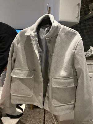 Ljusgrå overshirt med stora fickor - Snygg ljusgrå overshirt med raka linjer och två stora fickor framtill. Jackan har en klassisk krage och stängs med knappar. Materialet ser ut att vara ullblandning, vilket ger en mjuk och stilren känsla. Perfekt för lager-på-lager och trendig streetstyle.
