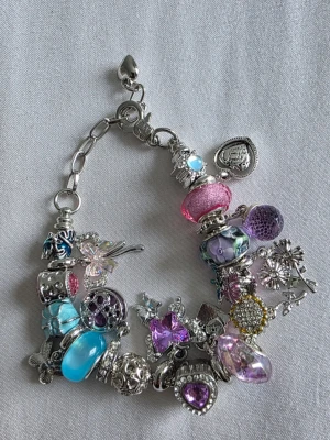 Silverfärgat armband med pärlor och berlocker - Armband i silverfärg med flera färgglada pärlor och berlocker i blått, rosa och lila. Några berlocker har hjärtform och glittrande detaljer. Materialet är metall och glas. Perfekt för dig som gillar unika accessoarer med mycket färg och detaljer.