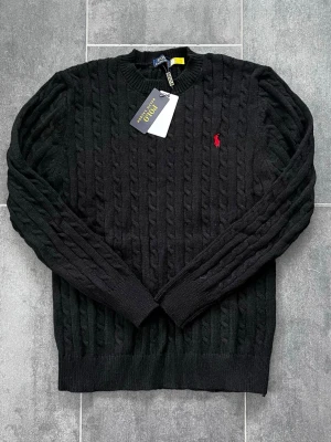Svart Polo Ralph Lauren - Kabelstickad svart tröja från Polo Ralph Lauren med rund halsringning och röd broderad logga på bröstet. Tröjan har ribbade muddar vid ärmslut och nederkant. Perfekt för dig som gillar klassisk och stilren design.