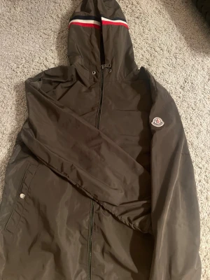 Moncler vindjacka med huva - Snygg mörkgrön vindjacka från Moncler med huva och dragkedja framtill. Jackan har långa ärmar, två fickor med tryckknappar och en vit-röd-blå rand på huvan. Materialet är tunt och vindtätt, perfekt för blåsiga dagar.