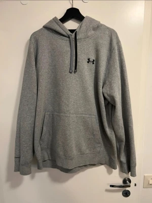 Grå hoodie från Under Armour - Snygg grå hoodie från Under Armour med svart logga på bröstet. Klassisk känguruficka framtill och justerbar dragsko i huvan. Tillverkad i mjukt sweatshirtmaterial som är perfekt för chill eller träning. Långärmad och med ribbade muddar.
