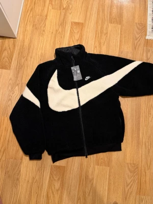 Nike svart fleece Jacka Big Swoosh Reversible  - Säljer en svart fleece cardiganjacka från Nike med stor vit Swoosh-logga över bröstet och ärmen. Jackan har dragkedj framtill, hög krage och muddar vid ärmslut och nederkant. Perfekt för dig som gillar sportig och trendig stil.