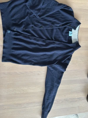 Mörkblå cashmere tröja från La chemise - Stilren svart kofta med rund halsringning och knäppning framtill. Tillverkad i ett tunt, mjukt material som ger en avslappnad passform. Ribbstickade muddar vid ärmslut och nederkant. Perfekt att slänga över en t-shirt eller linne.