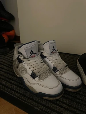 Air Jordan 4 White/Midnight Navy - Säljer ett par Air Jordan 4 i vitt läder med detaljer i mörkblått och grått. Skorna har meshpaneler på sidorna, klassisk Jumpman-logga på plösen och häl, samt en robust sula med synlig Air-enhet. Perfekta för dig som gillar retrostil och streetwear.