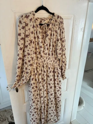 Blommig långklänning från Newbie - Romantisk långklänning från Newbie i ljusbeige med små vinröda blommor. Klänningen har långa ärmar, v-ringning med knytband och volangdetaljer vid halsen. Tillverkad i mjuk bomull, perfekt för sommarens alla tillfällen.