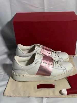 Valentino open skor - Snygga vita sneakers från Valentino med ett metallicrosa band över mitten. Skorna har klassisk låg profil, vita nitar på hälen och är tillverkade i läder. Perfekta för dig som vill ha en clean look med en twist. Levereras med originalkartong och extra skosnören.