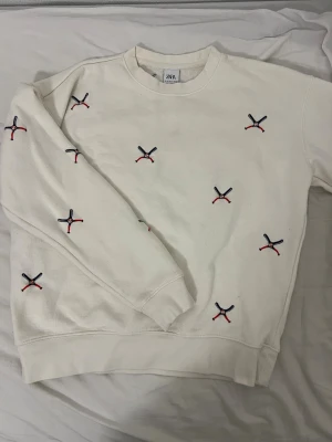 Vit sweatshirt med broderi från Zara - Vit sweatshirt från Zara med broderade detaljer i svart, rött och blått över hela tröjan. Rund halsringning och mjukt material på insidan. Perfekt för en avslappnad och trendig stil.