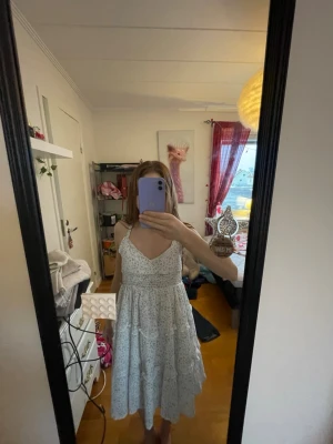 Vit blommig sommarklänning - Söt vit klänning med småblommigt mönster i blått och grönt. Klänningen har smala axelband, omlott-ringning och smock i midjan för en bekväm passform. Kjolen är volangig med dekorativa sömmar och lätt rynk. Perfekt för varma dagar 