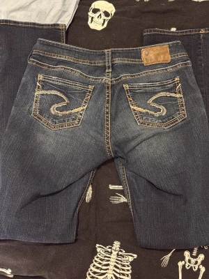 Mörkblå bootcut jeans från Silver - Säljer ett par mörkblå bootcut jeans från Silver Jeans Co. med slitningar och snygga kontrastsömmar. Bakfickorna har broderade detaljer i beige och jeansen har en klassisk femficksmodell. Materialet är denim i bomull med lätt stretch. säljer för att jag inte gillar lågmidjat💗