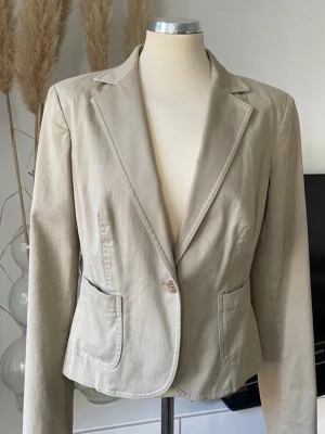 Beige figursydd kavajGANT - Stilren beige kavaj med figurnära passform och klassisk krage. Kavajen har två framfickor och stängs med en knapp framtill. Tillverkad i ett mjukt bomullsliknande material som ger en avslappnad men ändå dressad look.