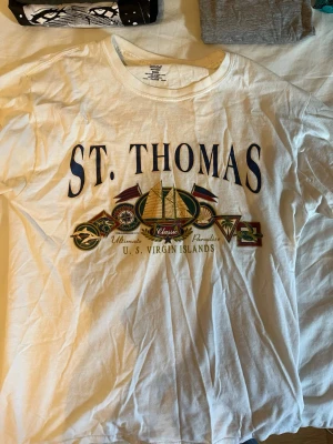 St. Thomas t-shirt med tryck - Vit t-shirt med stort St. Thomas-tryck framtill och färgglada detaljer som båt, flaggor och kompass. Klassisk rund hals och korta ärmar. Perfekt för dig som gillar semester vibes och vill sticka ut lite extra.