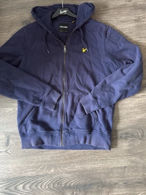 Marinblå hoodie från Lyle & Scott - Snygg marinblå hoodie med dragkedja från Lyle & Scott. Klassisk gul logga på bröstet, två fickor fram och justerbar huva med snören. Tillverkad i mjuk bomullsblandning för skön känsla. Perfekt för en avslappnad och stilren look.